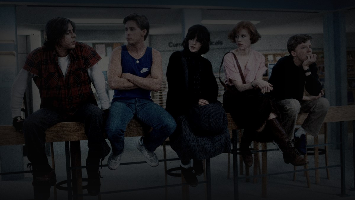 Still from The Breakfast Club (1985) — letterboxd og:image (a.ltrbxd.com/resized/sm/upload/6y/th/32/zi/breakfast-club-1200-1200-675-675-crop-000000.jpg); genuine production still, all five leads sitting together on the library table with the counselor books sign behind them. source luma 96.5, pre-darkened 0.75, baked at factor 0.45, lanczos to 1200x675, on-disk luma 23.6